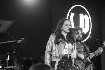 A black and white image of a woman singing into a microphone on stage, wearing a leather jacket and a printed shirt. A man in the background plays a bass guitar, and there's a circular logo with letters behind them.