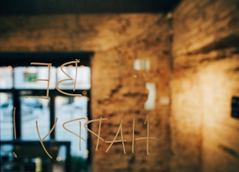 Words written in a semi-transparent manner on a window or glass surface. The background features an interior setting with blurred brick walls and large windows. The natural light creates a warm ambiance.