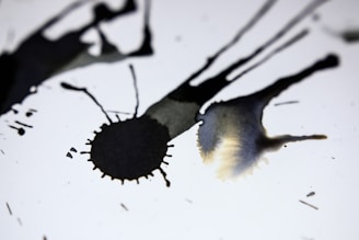 Dark ink splatters spread across a white background, creating abstract shapes and patterns. The ink varies in intensity and opacity, creating a dynamic contrast against the bright surface.