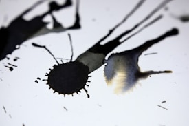 Dark ink splatters spread across a white background, creating abstract shapes and patterns. The ink varies in intensity and opacity, creating a dynamic contrast against the bright surface.