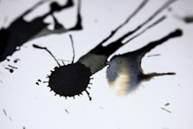 Dark ink splatters spread across a white background, creating abstract shapes and patterns. The ink varies in intensity and opacity, creating a dynamic contrast against the bright surface.