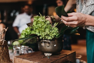A person is arranging green flowers and leaves in a vintage, brass pot on a wooden surface. The setting appears to be indoors with soft, warm lighting. Other people are blurred in the background, suggesting a social or workshop environment. Additional items include a woven basket, a glass measuring cup, and a pencil scattered on the table.