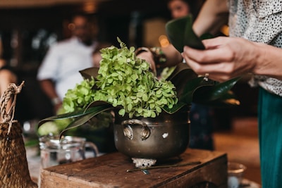A person is arranging green flowers and leaves in a vintage, brass pot on a wooden surface. The setting appears to be indoors with soft, warm lighting. Other people are blurred in the background, suggesting a social or workshop environment. Additional items include a woven basket, a glass measuring cup, and a pencil scattered on the table.