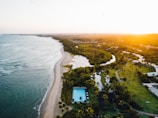 Aerial view of a coastal landscape featuring a long sandy beach with gentle waves, a swimming pool surrounded by palm trees, and a river that winds through lush greenery. The sun is setting in the background, casting a warm golden light over the scene. There are scattered buildings and roads visible among the trees.