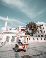 A street vendor cart with an orange and white striped canopy is stationed on a paved path, selling baked goods. The vendor is standing behind the cart, and the scene takes place in front of a historic building with domes and minarets. The sky is clear with a few clouds.