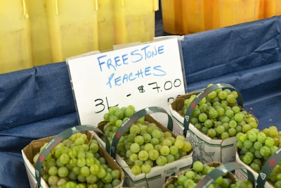 Baskets of green grapes are displayed in front of a sign that reads 'Free Stone Peaches 3L - $7.00'. The setup is on a blue cloth, and there are yellow plastic bins in the background.