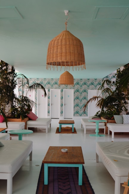 A stylish and tropical-themed interior space decorated with white lounge chairs and wooden tables. Woven light fixtures hang from the ceiling, and potted plants with lush green leaves are scattered around. The walls are decorated with a leafy wallpaper that adds to the tropical vibe. Cushions in pastel hues of pink and green complement the serene atmosphere.