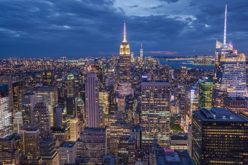 A vibrant cityscape at dusk, featuring a skyline filled with illuminated skyscrapers. The sky is a mix of deep blues and hints of sunset pink, reflecting off the buildings. The city lights create a glowing atmosphere as they contrast with the darkening sky.