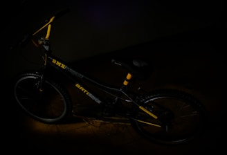 A BMX bicycle with yellow accents is placed in a dimly lit environment. The frame of the bike is primarily black, featuring a BMX logo and other graphics. The handlebars are partially visible, and the tires have a rugged texture.
