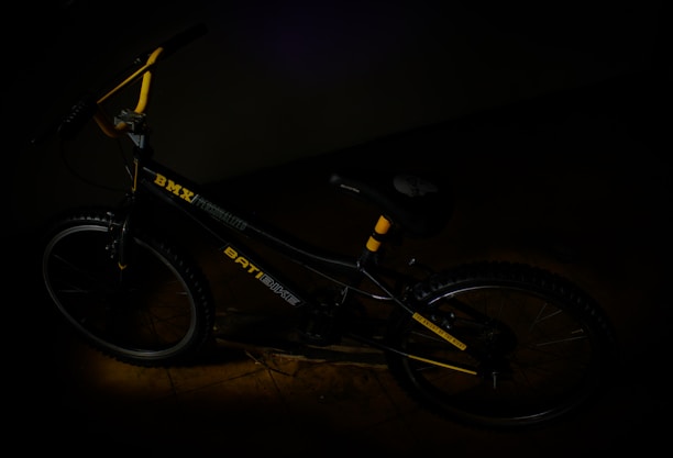 A BMX bicycle with yellow accents is placed in a dimly lit environment. The frame of the bike is primarily black, featuring a BMX logo and other graphics. The handlebars are partially visible, and the tires have a rugged texture.