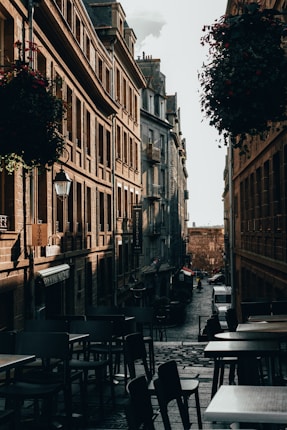 A narrow cobblestone street flanked by tall, historic buildings bathed in soft, warm light. Outdoor caf&eacute; tables and chairs line the foreground, while hanging flower baskets add a touch of greenery. In the distance, a person walks down the street past parked cars, conveying a quiet, early morning or late afternoon atmosphere.