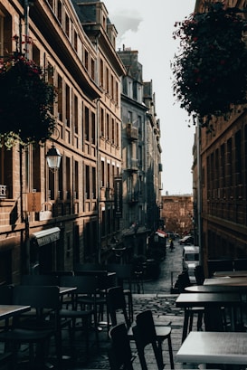 A narrow cobblestone street flanked by tall, historic buildings bathed in soft, warm light. Outdoor caf&eacute; tables and chairs line the foreground, while hanging flower baskets add a touch of greenery. In the distance, a person walks down the street past parked cars, conveying a quiet, early morning or late afternoon atmosphere.