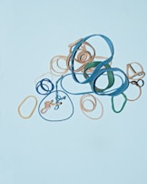 A variety of rubber bands in different colors, including blue, beige, green, and yellow, are arranged on a light blue background. The rubber bands are tangled and overlapping, creating an abstract pattern.