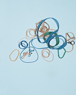 A variety of rubber bands in different colors, including blue, beige, green, and yellow, are arranged on a light blue background. The rubber bands are tangled and overlapping, creating an abstract pattern.