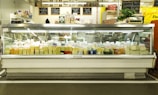 A large display case filled with various types of cheese, each labeled with tags. Above the case, there are signs describing the products and prices. The setting appears to be a cheese shop with a variety of cheeses in different shapes and colors.