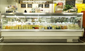 A large display case filled with various types of cheese, each labeled with tags. Above the case, there are signs describing the products and prices. The setting appears to be a cheese shop with a variety of cheeses in different shapes and colors.
