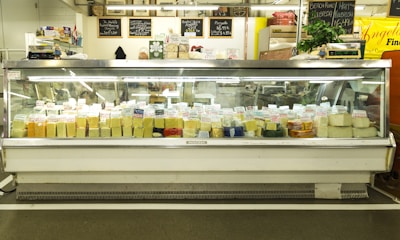 A large display case filled with various types of cheese, each labeled with tags. Above the case, there are signs describing the products and prices. The setting appears to be a cheese shop with a variety of cheeses in different shapes and colors.