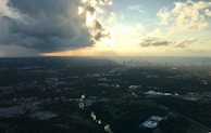 A dramatic aerial view of a cityscape with dark, looming storm clouds on the left and clear skies on the right as sunlight breaks through, creating a stark contrast. The city below is partially shrouded in shadow, emphasizing the mixed weather conditions.