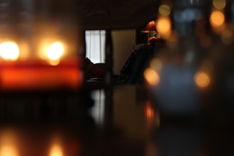 A dimly lit room with a cozy and intimate atmosphere. Soft focus highlights a glowing table lamp and blurred reflections, creating a warm ambiance. The candles emit a gentle light, casting a serene glow.