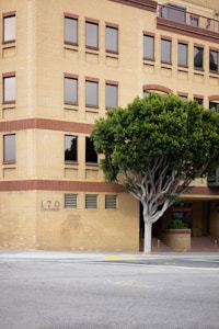 A multi-story brick building with evenly spaced windows, adjacent to a sidewalk and a large leafy tree in the foreground. The building has a blend of tan and red bricks and features arched entranceways with a partially visible sign that reads 'Urgent Care.' There are no people on the street, which appears quiet and empty.