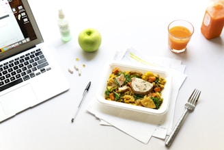 A tidy workspace includes a laptop displaying an email interface, a black pen, and several white capsules on the left. A green apple and a spray bottle sit nearby. In the center, a container of healthy food with chicken and vegetables is placed on top of scattered papers. To the right, a glass of orange juice and an orange juice bottle are set on the table, next to a metal fork.