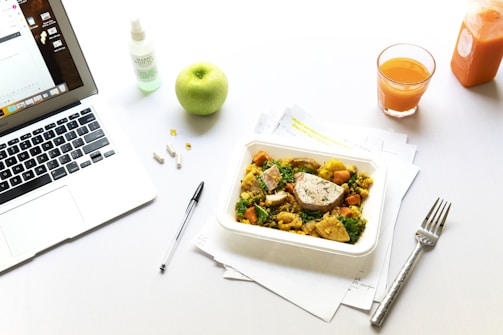 A tidy workspace includes a laptop displaying an email interface, a black pen, and several white capsules on the left. A green apple and a spray bottle sit nearby. In the center, a container of healthy food with chicken and vegetables is placed on top of scattered papers. To the right, a glass of orange juice and an orange juice bottle are set on the table, next to a metal fork.
