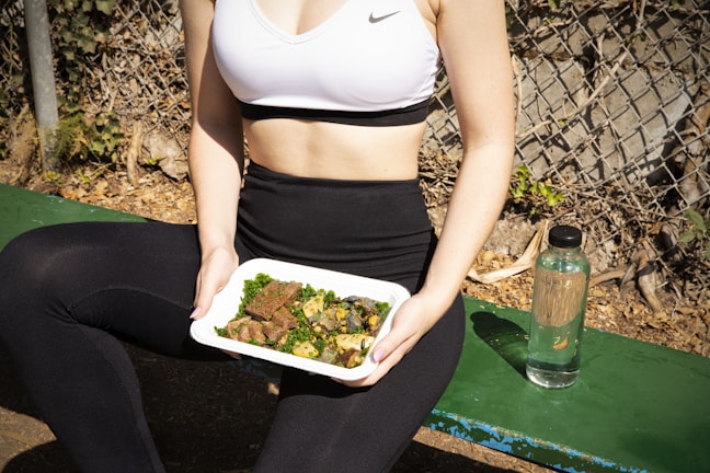 A person wearing a white sports bra and black leggings is sitting on a green bench beside a chain-link fence. They are holding a rectangular food container filled with a meal consisting of greens and cooked meat. A clear bottle with a black cap is placed next to them on the bench. The setting appears to be outdoors with a natural background of leaves and twigs.