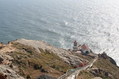 A lighthouse with an attached house is situated on a rocky cliff overlooking a vast, sparkling ocean. A staircase leads down to the lighthouse and house from the higher cliff edge. The scene is bathed in sunlight, creating reflections on the water.
