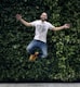 man wearing t-shirt and jeans jumpshot in front of a green hedge