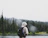Close-up of a person wearing a forest green hoodie with gold accents, standing next to a floatplane on a misty Alaskan lake.