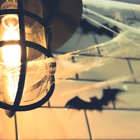 An industrial-style light fixture is surrounded by artificial cobwebs, creating a spooky atmosphere. A silhouette of a bat is visible in the background against tiled walls, enhancing the Halloween theme.