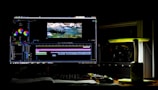 A dark room with a computer setup displaying video editing software on the monitor. The screen shows a video timeline and some color correction panels. Beside the monitor is a lamp casting a soft greenish-yellow light. Various cables and a mouse are placed on the desk.