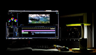 A dark room with a computer setup displaying video editing software on the monitor. The screen shows a video timeline and some color correction panels. Beside the monitor is a lamp casting a soft greenish-yellow light. Various cables and a mouse are placed on the desk.