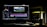 A dark room with a computer setup displaying video editing software on the monitor. The screen shows a video timeline and some color correction panels. Beside the monitor is a lamp casting a soft greenish-yellow light. Various cables and a mouse are placed on the desk.