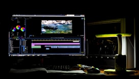 A dark room with a computer setup displaying video editing software on the monitor. The screen shows a video timeline and some color correction panels. Beside the monitor is a lamp casting a soft greenish-yellow light. Various cables and a mouse are placed on the desk.