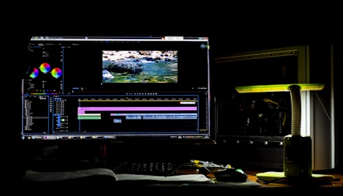 A dark room with a computer setup displaying video editing software on the monitor. The screen shows a video timeline and some color correction panels. Beside the monitor is a lamp casting a soft greenish-yellow light. Various cables and a mouse are placed on the desk.