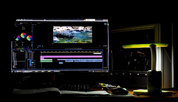 A dark room with a computer setup displaying video editing software on the monitor. The screen shows a video timeline and some color correction panels. Beside the monitor is a lamp casting a soft greenish-yellow light. Various cables and a mouse are placed on the desk.