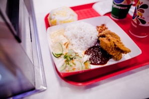 A red tray holds a plate of food featuring a serving of white rice, crispy fried chicken, sambal sauce, cucumber slices, and fried anchovies. There is also a wrapped burger and two beverages in colorful cups nearby.
