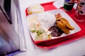 A red tray holds a plate of food featuring a serving of white rice, crispy fried chicken, sambal sauce, cucumber slices, and fried anchovies. There is also a wrapped burger and two beverages in colorful cups nearby.