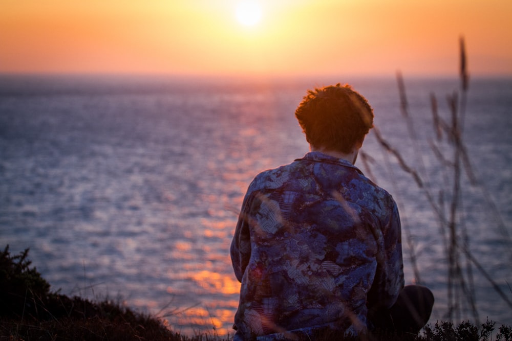 man wearing black-and-blue floral dress shirt sitting on grass near body of water during golden hour