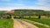 A rural landscape featuring a dirt road leading through lush green fields. In the background, there is a cluster of houses surrounded by fields of various crops, including a distinct yellow patch likely representing rapeseed or mustard fields. Trees and hedges are interspersed throughout the landscape, and the scene is set under a clear blue sky.