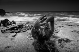 A person lies on a sandy beach reaching out towards the camera. The waves from the ocean can be seen in the background, crashing onto the shore. Various items are scattered on a blanket, including a backpack and camera equipment. The atmosphere is relaxed and carefree, suggesting a day of leisure at the beach.