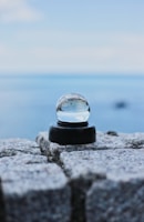 A crystal ball is placed on top of a stone surface with a blurred seascape in the background. The sky is clear with soft clouds, and the water appears still and serene.