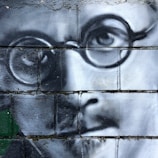 A detailed black and white mural painted on a brick wall depicting the close-up of a man's face wearing round glasses. The mural is rendered with realistic shading and contouring, focusing on the eye and part of the nose and mouth.