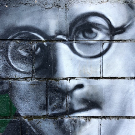 A detailed black and white mural painted on a brick wall depicting the close-up of a man's face wearing round glasses. The mural is rendered with realistic shading and contouring, focusing on the eye and part of the nose and mouth.