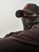 A person wearing a dark cap with orange lettering and a face covering, possibly to protect against cold weather. The cap has a curved brim, and the clothing appears to be layered.