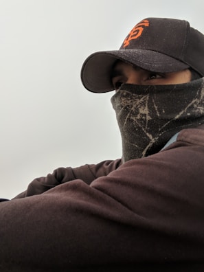 A person wearing a dark cap with orange lettering and a face covering, possibly to protect against cold weather. The cap has a curved brim, and the clothing appears to be layered.