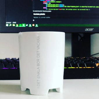 A white mug with Swedish text printed on it is placed in front of a computer monitor displaying a code editor. The keyboard in front of the monitor has colorful backlighting.