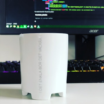 A white mug with Swedish text printed on it is placed in front of a computer monitor displaying a code editor. The keyboard in front of the monitor has colorful backlighting.