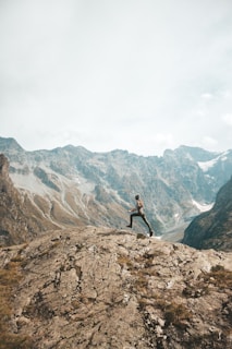 A person jumps energetically over a rocky terrain with a backdrop of towering mountains under a cloudy sky. The scene conveys a sense of freedom and adventure, with rugged geological formations and patches of grass accentuating the natural landscape.